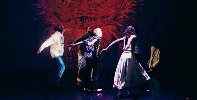 Four dancers perform in front of a red orb projected behind them.