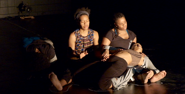 In a spotlight, three women lay together. One is sprawled over two them and potentially unconscious.