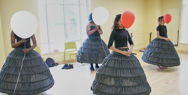 Four women in black dresses each hold a single red or white balloon in front of their faces in a dance studio.