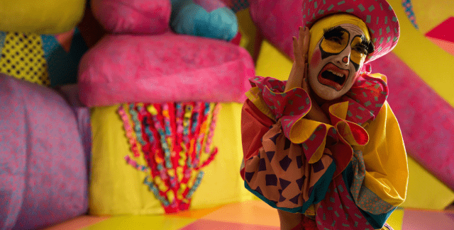 A drag queen dressed like a clown stands in front of a colorful background.