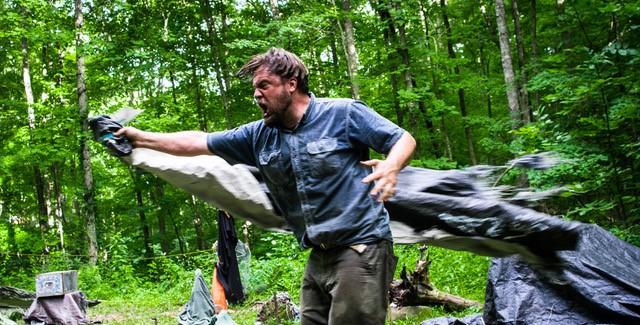 In the woods, a man charges forward with something beneath a coat or fabric.