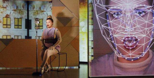 A woman on a stage sits next to a live video of herself that is close on her face and has lines mapped to parts of her face.