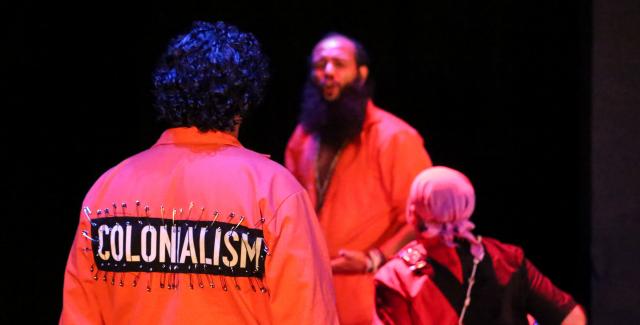 A man in an orange jumpsuit has pinned the word "Colonialism" to the back of it.