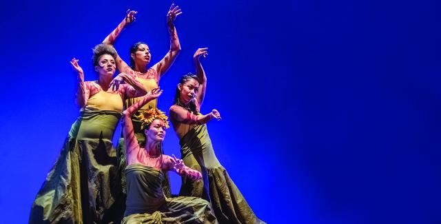 Four women in a diamond formation dance in front of a blue backdrop.