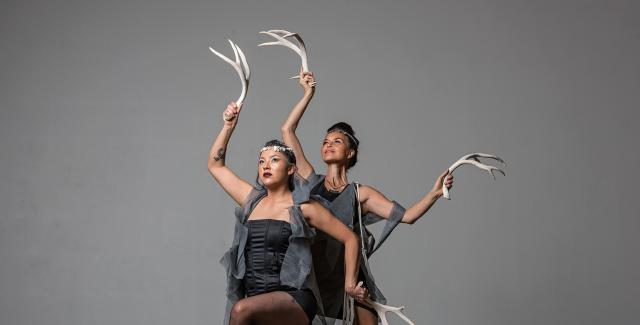 Two females dance in front of a gray backdrop with antlers in their hands.