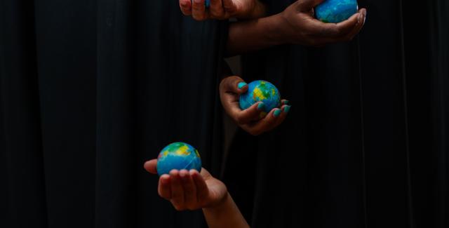 Five hands poke out from a set of black curtains, each one holds a miniature globe.