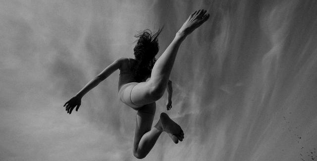 Black and white image, from underneath a person either leaps or is swimming. Stark contrast with their legs bent back and their hands reaching forward.