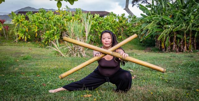 A Hawaiian woman lays low to the ground and holds up two bamboo sticks that create an X.