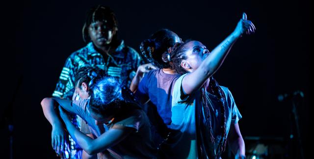 On a dark stage, with spotlights, four dancers of color look and point to the sky. They wear or appear in the light to wear blues and purples.