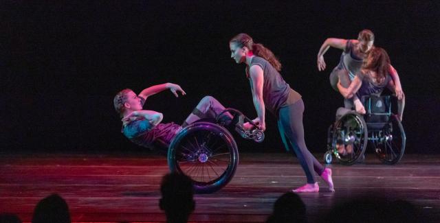 A standing dancer pushes a dancer on wheels by his feet.