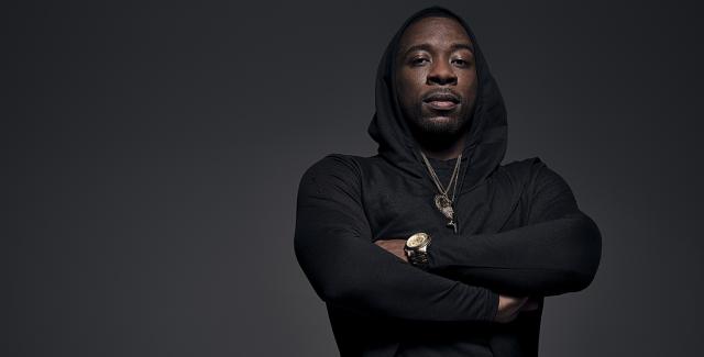 A black man crosses his arms. He poses in front of a gray backdrop and wears a black hoodie.