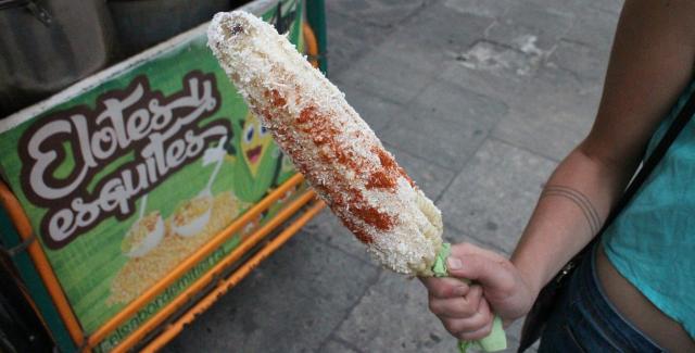 A woman holds an elote in front of a cart that also says, "Elotes."