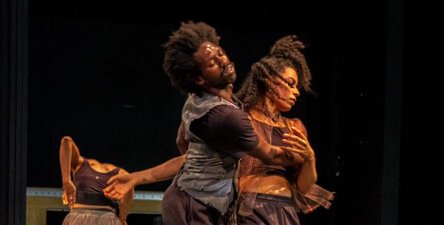 On a stage, A Black man leads a Black woman in a waltz-like dance.