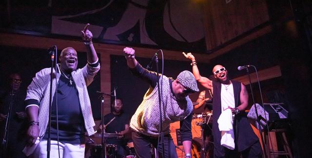 Three Black men dance and point. One bends over. A band is behind them. 