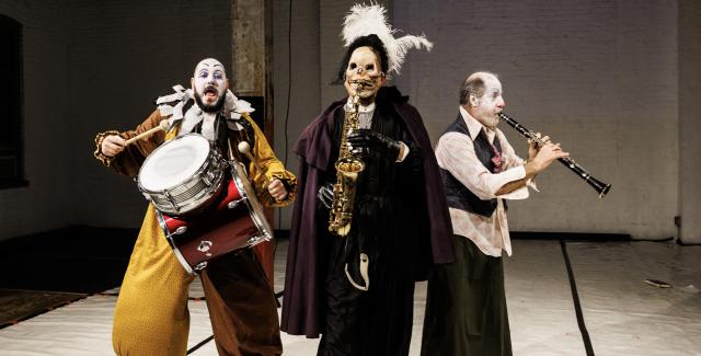 On a stage, three men in clown gear that is in sad hues and one of them has a skull mask on..