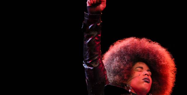 A Black girl, with an Afro, holds up her fist in a spotlight.