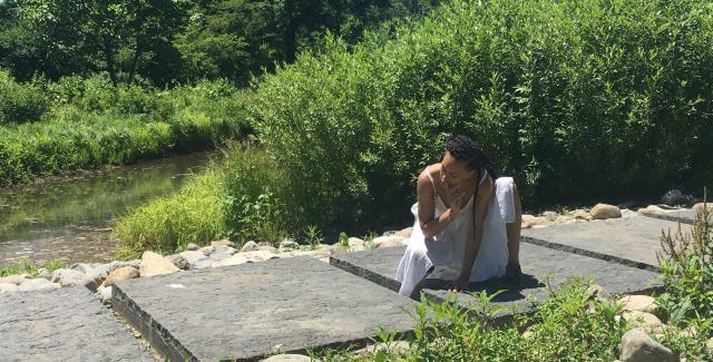 Along a stone path, a woman of color, dressed in white, crouches on one knee.
