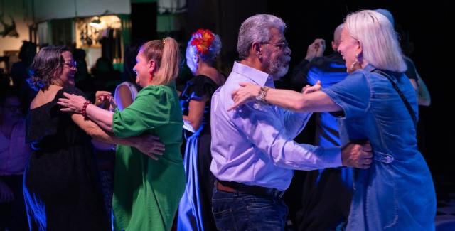 In the foreground, an elderly heterosexual couple dances. Behind them, younger couples of various combinations dance.