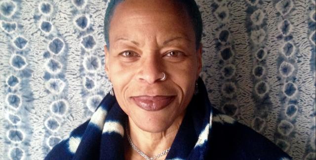 Ife wears blue and poses with a blue package on her head in front of a wall with a blue floral wallpaper.