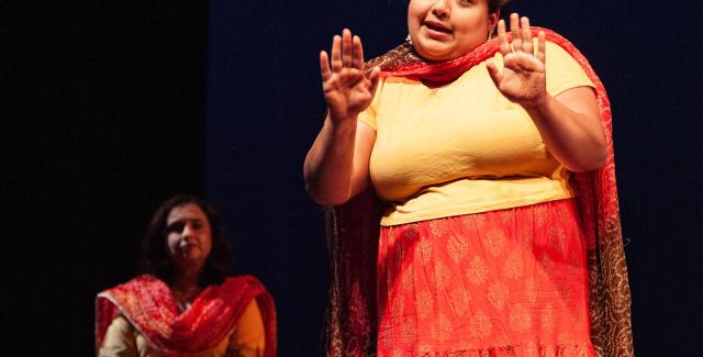Two brown women are on stage. They wear red and yellow outfits, including wraps that lay by their necks and across their bodies. The woman in the foreground holds her hands up.