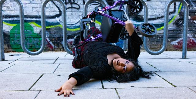 a photo of Vanessa, who is a Chicana woman with caramel skin is lying down on the concrete floor with her arm reaching out as she is looking back towards the camera. Her walker Pluto is balancing on top of her. She is wearing a gothic long sleeve blouse with a flowered black and red skirt. In the background is a looping metal bicycle stand with a graffiti filled wall.]