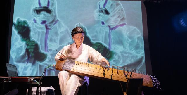Jin Hi plays the electric komungo, wearing all white in front of a projection of folks in hazmat suits.