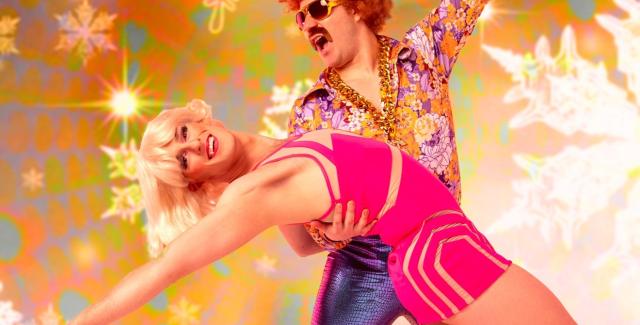 Two colorfully dressed performers dance together in front of a background of gold and snowflakes.