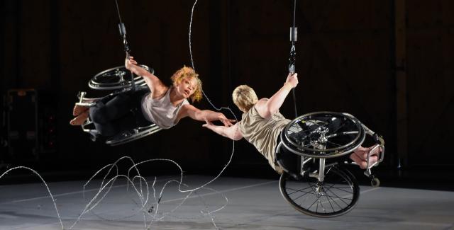 Alice, a dancer with light brown skin and short curly hair, and Laurel, a dancer with pale skin and close-cropped blonde hair, hang from the ceiling. They each have one hand on a cable as they reach for each other with the other. They are nearly horizontal and their legs and feet face out. Silver barbed wire stretches between and beneath them, against the black backdrop.