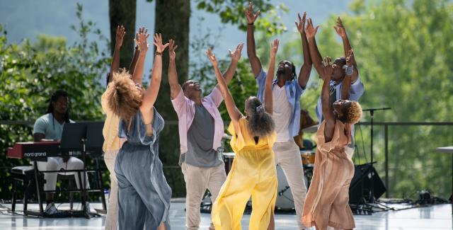 At an outdoor space, six Black dancers, in jumpsuits and khakis, hold their hands up to the sky.