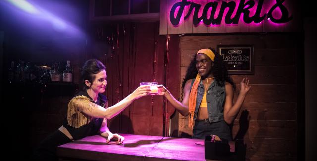 Two women stand by a bar that says "Frank's" in neon behind them,