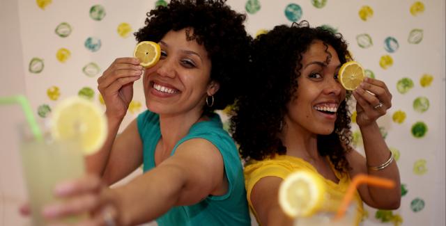 Elisa and Silvia hold a slice of lemon in front of their eye and out of focus in front of them, they also hold a glass of lemonade with a staw.