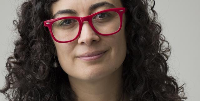 Lina, in red glasses and a floral shirt, smirks in front of a grey wall.