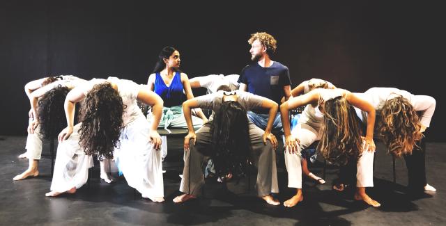 Two dancers look at each other while surrounded by female dancers with their heads down and their hands on their knees.