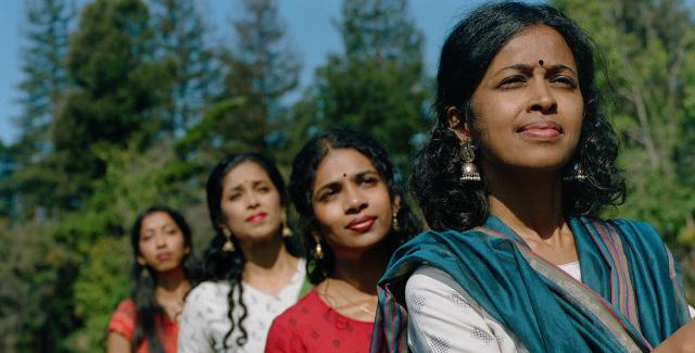 Four west Asian women wear saris in a river. They all face the same direction, toward the camera, and pose in a line.