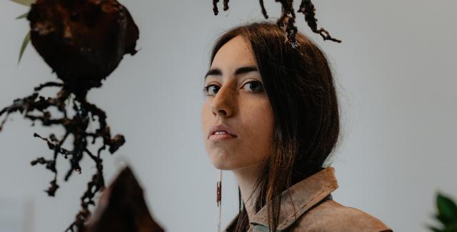 Mali has long brown hair. She wears a khaki shirt and a long earing. She poses next to earthy sculptures that hang from the ceiling.