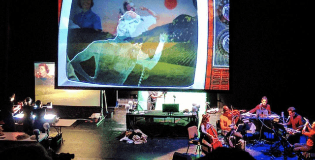 Puppeteers and an orchestra are on either side of a projection of a TV that shows a character running.