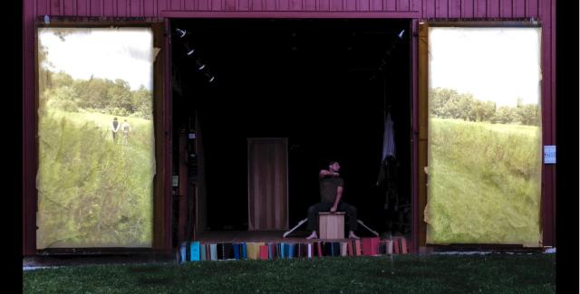 Outside, a man performs in a barn, where the doors have projections of fields on them.