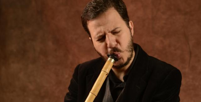 Mehmet plays a reed instrument in front of a brown background.