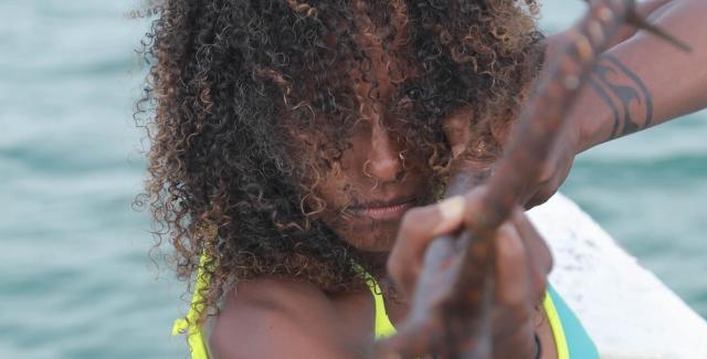 A Black woman, in a bikini and with shoulder length curly hair, aims an anchor or sling shot or some kind of metal piece at the camera.