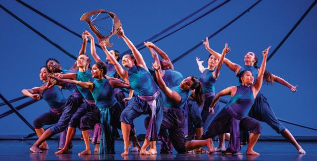 On a stage, a troupe of dancers wear blue and purple in front of a blue backdrop.