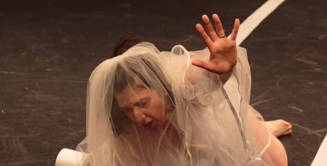 Sara lays on the ground, wearing a bridal veil, and raises her hand up as if to reach.