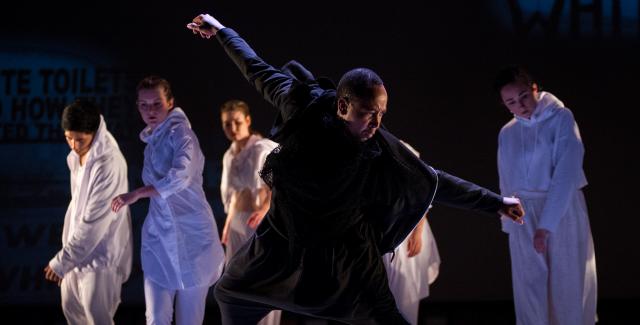A man in black dances in the foreground while dancers in white dance in the background.