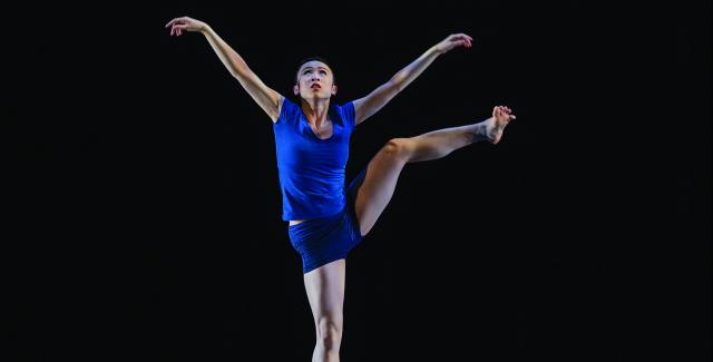 A woman in blue in front of a black backdrop raises her left leg and both arms.