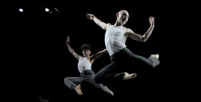Two male dancers leap behind three female dancers who are unconscious next to an apple with a bite taken out of it.