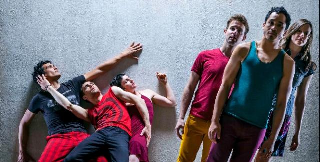 Three dancers wearing bright colors lean against a wall while three more lean to the left in the foreground.