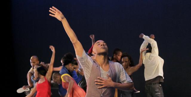 A man in a vest throws his arm up in the foreground and in the background a group of dancers move in a circle.