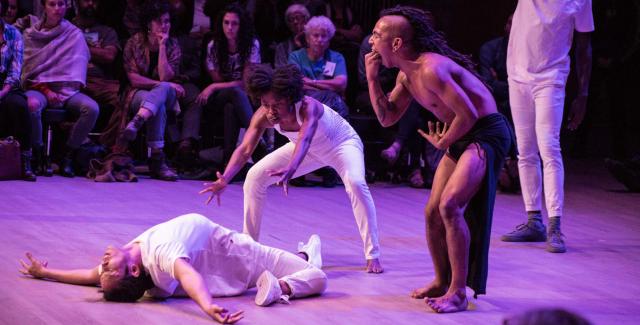 A man dressed in Native American garb pretends to vomit on two women who are dancing in white and look to be exorcising a demon from one of the women