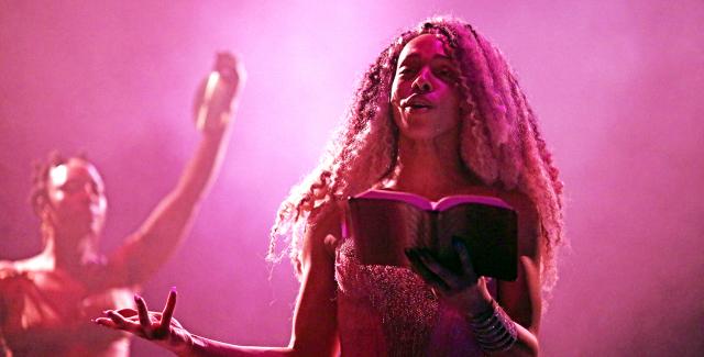 In front of a dancer and in pink light, a Black woman reads from a book.