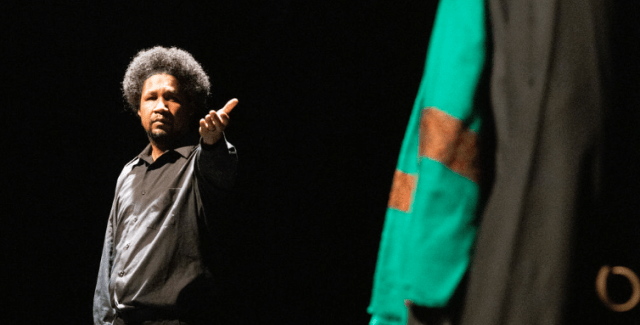 On a stage and under spotlights, a Black man, in all black, holds his hand out to a brown man in a green scarf and black robe.