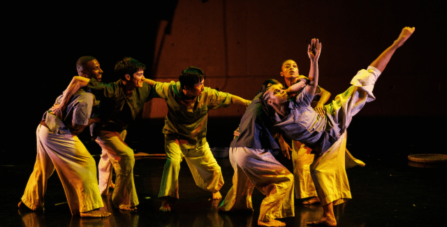 A group of six performers are on a stage with their arms slung over each others shoulders. The one on the far right raises a leg and arm towards the sky.